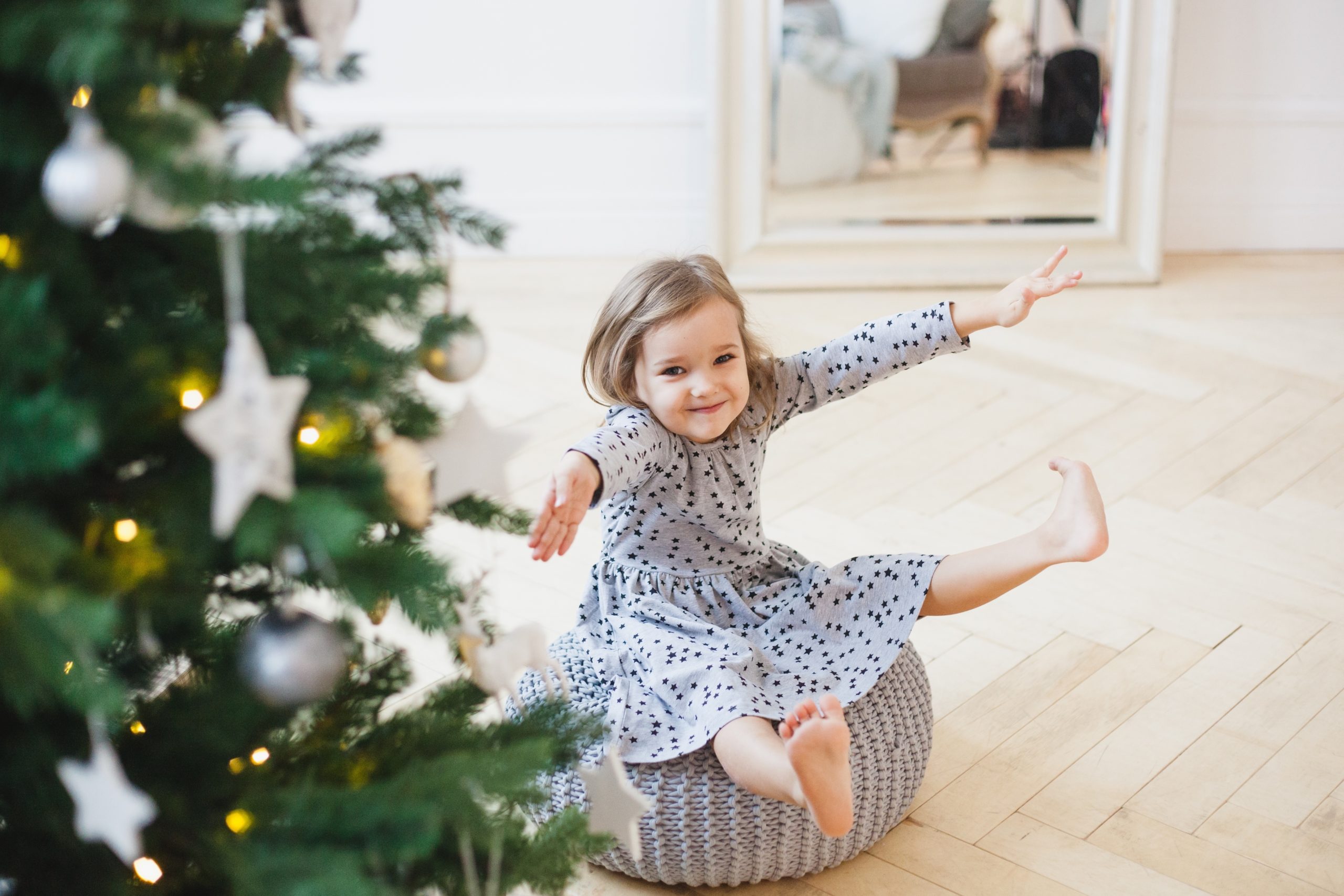 excited little girl sitting on ottoman pouf next to christmas tree-family secret santa