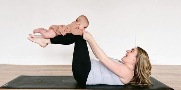 mom doing reclined boat pose while holding baby's hand - yoga with baby