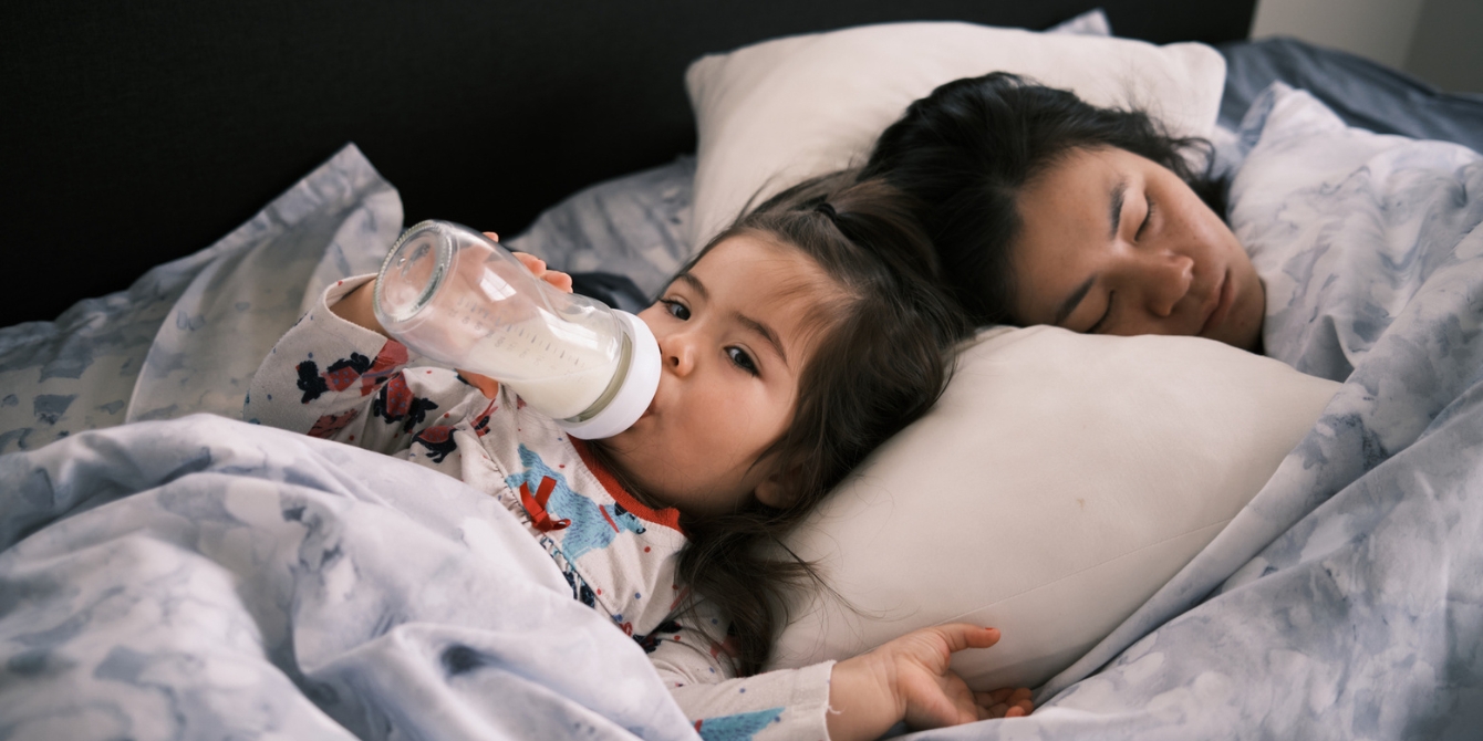 toddler girl drinks milk next to mom in bed