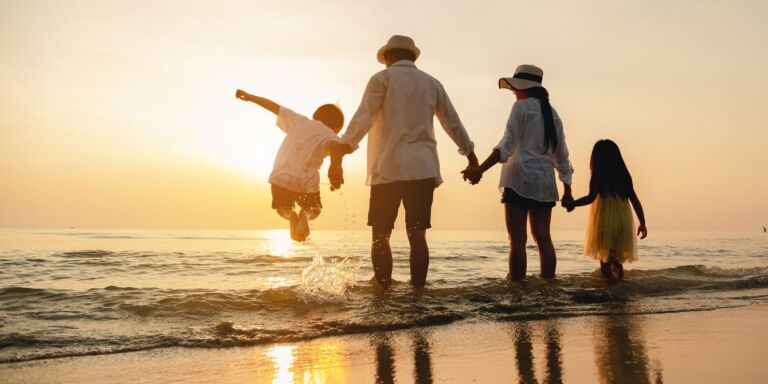 mom, dad and two kids holding hands standing at the beach during sunset and one child is jumping